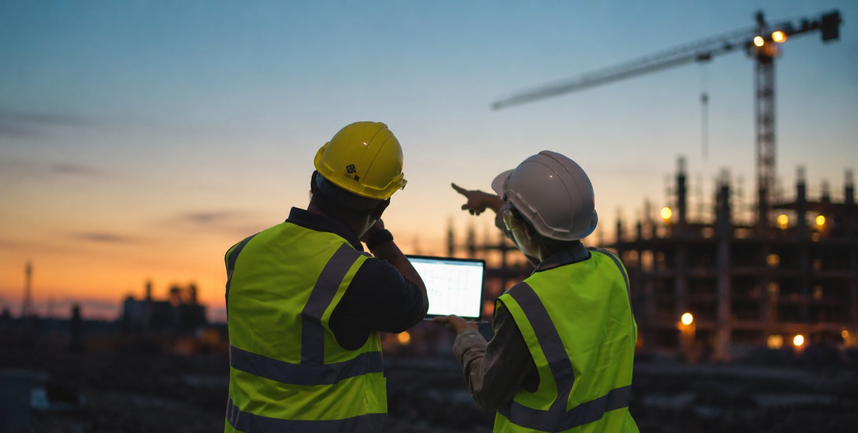 Two site supervisors in hard hats reviewing plans on a tablet at a construction site at dusk